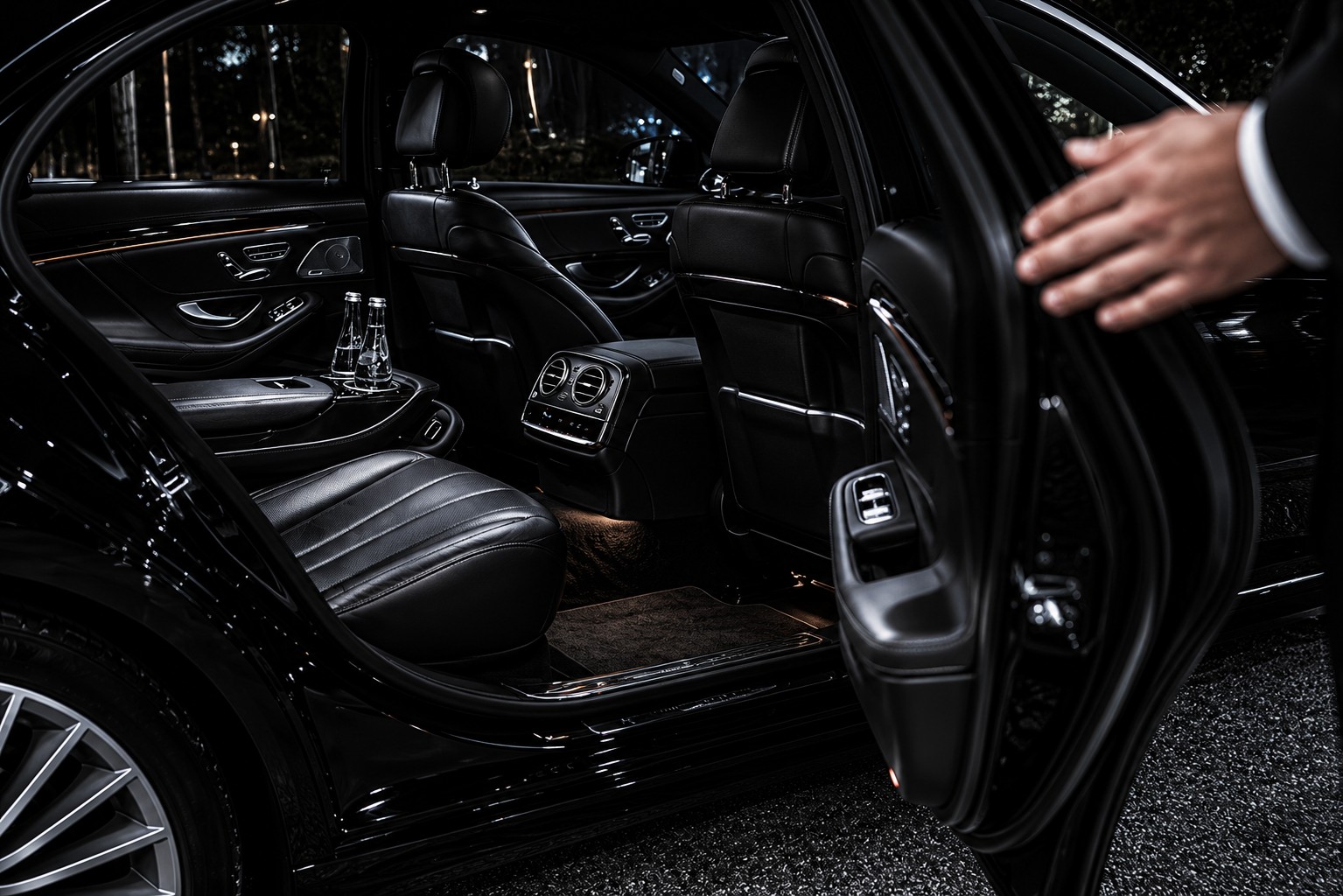 Habitacle arrière d'une Mercedes Classe S noire de la Maison Vehira : sièges en cuir, eau fraîche, portière tenue ouverte par le chauffeur.